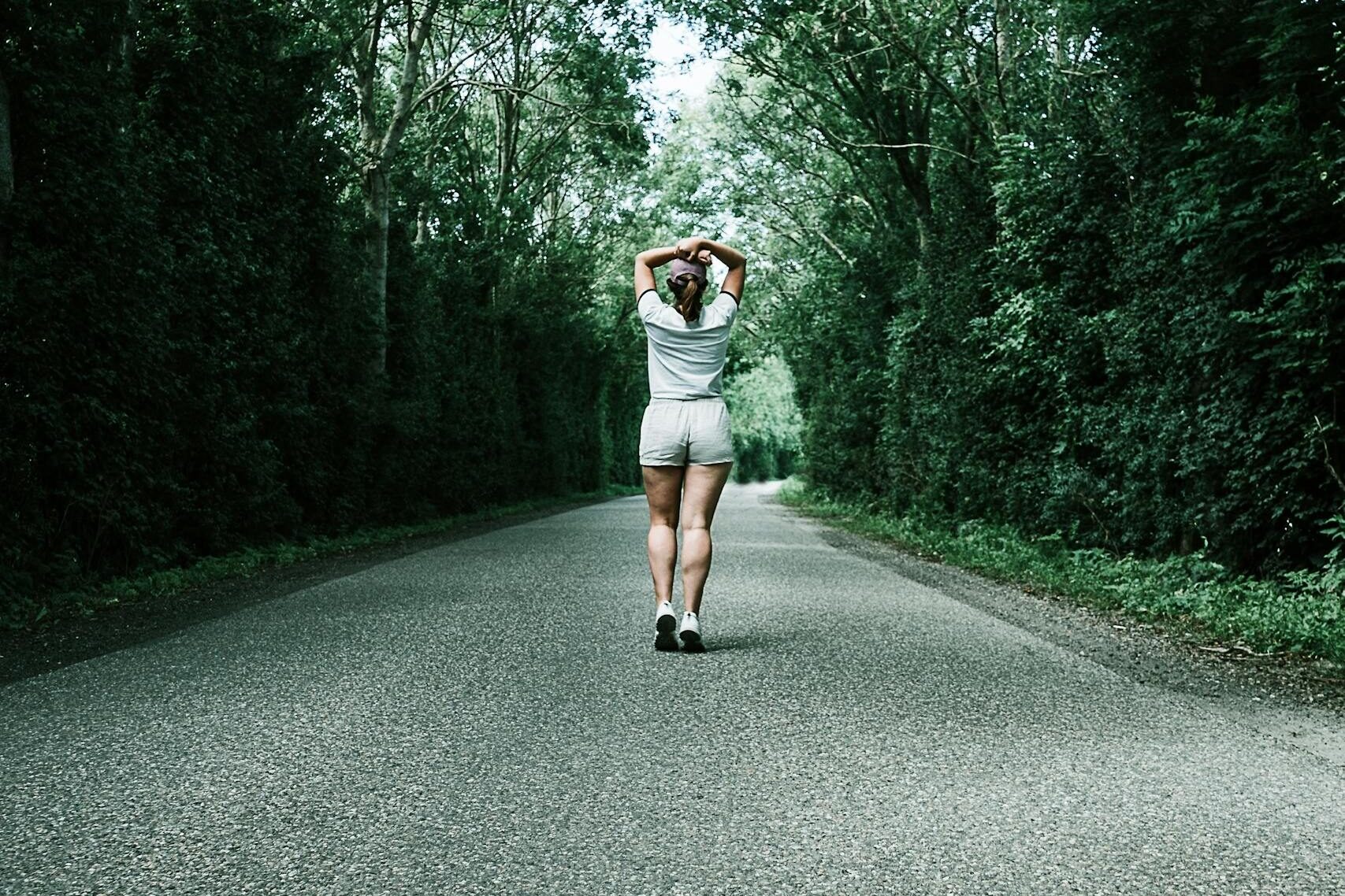 A woman jogs down a tree-lined road, embodying fitness and nature in a serene summer setting.
